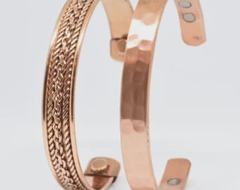May include: Two copper-colored metal cuff bracelets. One bracelet features a central braided rope pattern with smooth edges. The second bracelet has a hammered, dimpled texture. Both open-ended cuffs have small circular magnets on the inner ends.