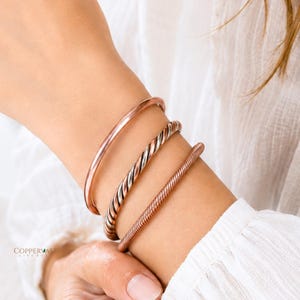 May include: A set of three copper-toned metal bracelets worn on the wrist. The set includes one smooth, polished cuff, one twisted rope-style bracelet, and one textured, ribbed cuff. The text COPPERVAST LIVE PURE is visible in the bottom corner.