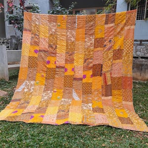 Op de afbeelding: Een grote patchwork quilt met warme kleuren, waaronder tinten geel, oranje en bruin. De quilt bestaat uit talrijke rechthoekige en vierkante stukken stof, sommige met bloempatronen en andere met effen kleuren. Paarse accenten zijn verspreid.