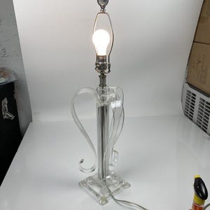 Op de afbeelding: Een heldere acryl tafellamp met een vierkante basis en een decoratief, gebogen ontwerp. De lamp heeft een zilverkleurige metalen steel en een gloeilamp met een donkergrijze eindkap. De lamp is aangesloten en brandt.