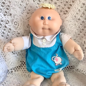 Puede incluir: Una muñeca Cabbage Patch Kid vintage con un cuerpo suave. La muñeca tiene una cara redonda, ojos azules y un mechón de pelo rubio. Lleva un pelele azul con cuello blanco y un pequeño diseño de elefante bordado. Un juguete coleccionable clásico.