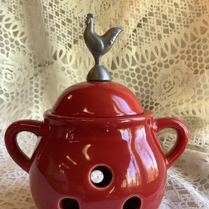 May include: A red ceramic garlic keeper with a lid and two handles. The lid is topped with a silver rooster figurine. The keeper has several round holes and is designed to store garlic. The keeper is on a white lace tablecloth.