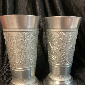 Könnte beinhalten: Zwei silberne Zinnbecher mit einem verzierten Blumenmuster. Die Becher haben einen breiten Boden und eine schmale Oberseite.