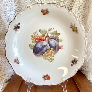May include: White porcelain serving plate with a scalloped edge and gold trim. The plate features a central illustration of plums, currants, and other fruits. Small fruit clusters are scattered around the rim.