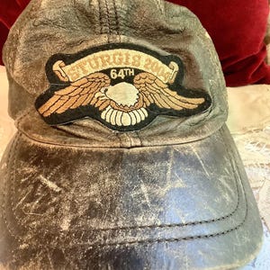 Peut inclure: Casquette de baseball en cuir gris vieilli avec un écusson brodé. L'écusson porte l'inscription "STURGIS 2004 64TH" et présente un motif d'aigle. La casquette a une visière incurvée et des coutures visibles.