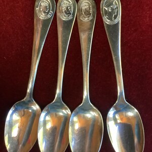 Puede incluir: Cuatro cucharas plateadas con cuencos ovalados y mangos largos. Cada mango de la cuchara presenta un retrato grabado detallado. Las cucharas se exhiben sobre una superficie de color rojo oscuro, que refleja la luz.