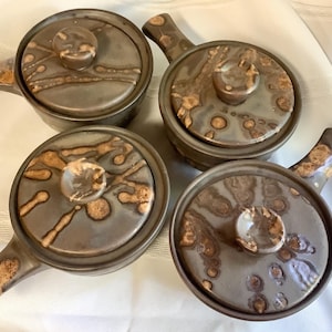 May include: A set of four round ceramic pots with lids and side handles. Each pot features a smooth, dark brown glaze with a decorative, lighter tan drip pattern across the lids and handles, creating a consistent look across all four pieces.