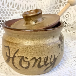 May include: A ceramic honey pot in shades of tan and brown, with a lid and a wooden honey dipper. The word "Honey" is written in brown script on the front. A floral design is on the right. The lid is slightly open.