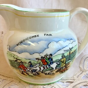 May include: Cream ceramic jug with a handle and spout, featuring a hand-painted scene of a fair with figures, animals, and the text "Widdicombe Fair". The rim is trimmed with green and yellow.