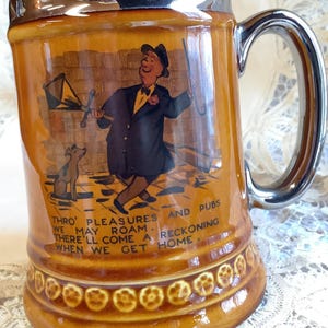 May include: A brown ceramic beer stein with a silver rim and handle. It features a cartoon illustration of a man in a suit and hat walking with a dog. The text on the stein reads "Thro' pleasures and pubs we may roam. There'll come a reckoning when we get home."