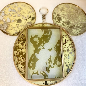 May include: A Mickey Mouse-shaped picture frame with a gold-coloured metal frame and a rectangular photo area. The frame has two large, round ears with a yellow and speckled design. The photo area features a silhouette of Mickey Mouse.
