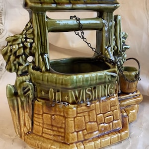 Può includere: Un vaso da fiori in ceramica verde e marrone a forma di pozzo dei desideri con una catena e un secchio. Il pozzo ha una base di mattoni e le parole "Oh Wishing" scritte sul davanti.