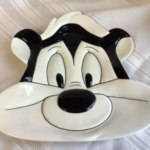 以下が含まれることがあります： 漫画のスカンクの顔の形をした白いセラミックプレート。スカンクは黒い毛皮、黒い鼻、黒い目を持っています。