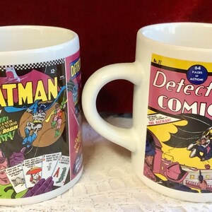 May include: Two white ceramic mugs with Batman comic book designs. One features "Batman" in yellow, the other "Detective Comics." Both mugs have black, pink, and yellow comic book graphics.