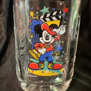 Puede incluir: Un vaso de vidrio transparente con una imagen colorida de Mickey Mouse vestido como director de cine. La imagen incluye una claqueta, una cámara y confeti. El vaso tiene el texto "Disney" y "2000" impreso.