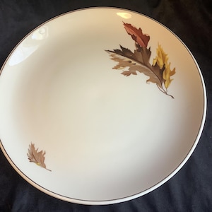 May include: A white ceramic dinner plate with a gold rim and a design of brown, yellow, and red leaves.