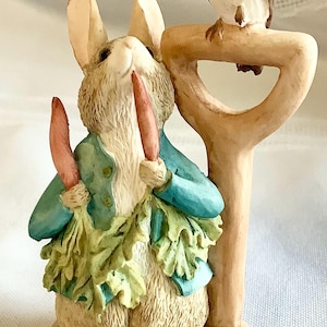 Può includere: Statuetta dettagliata di un coniglio che tiene due carote, indossa una giacca blu e una gonna verde a foglie. Un uccellino è appollaiato su una pala di legno. La statuetta è dipinta con colori naturali e ha un'estetica fiabesca.