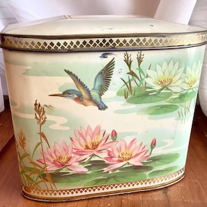 May include: Vintage metal storage tin with a cream lid and gold trim. The tin features a painted scene of a kingfisher in flight above pink water lilies and lily pads, set against a pale green background.