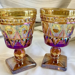 May include: Two iridescent glass goblets with a square base and a round stem. The goblets have a gold rim and a decorative pattern with purple and gold hues. The glasses are likely vintage or antique.