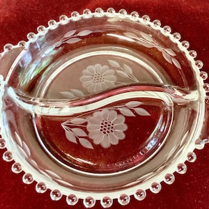 Puede incluir: Fuente de servir dividida de vidrio transparente con diseños florales grabados. El plato tiene un borde con cuentas y dos asas. El diseño floral incluye una flor grande y hojas. El plato está sobre una superficie roja.