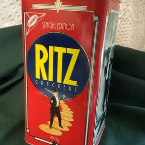 Puede incluir: Una lata rectangular roja de Ritz Crackers Edición Especial con tapa plateada. La lata presenta el logotipo de Ritz en amarillo y azul, un hombre con esmoquin caminando sobre galletas y el texto "Christie's". La lata contiene 450g de galletas.