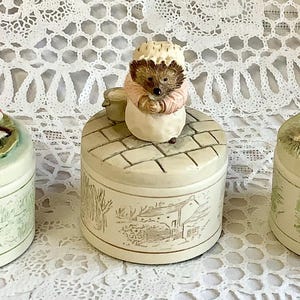 Puede incluir: Tres cajas de baratijas decorativas de cerámica con figuras de animales. Las figuras incluyen una rana, un erizo y un ganso. Las cajas son de color crema con un diseño de paisaje verde alrededor de la base.