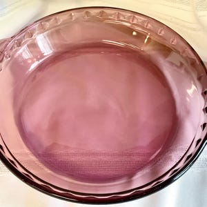 Puede incluir: Un molde para tarta redondo de vidrio transparente, de color púrpura claro. El plato tiene un borde decorativo con una serie de formas triangulares y dos asas. La base del plato tiene un patrón texturizado.