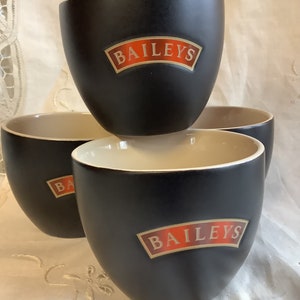 May include: Set of four black ceramic mugs with a cream coloured rim. Each mug features the Bailey's logo in gold lettering on a red banner.