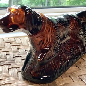 Puede incluir: Una figura de perro de cristal marrón con detalles en negro. El perro está en posición sentada, con la cabeza girada hacia la izquierda. La figura tiene un acabado brillante y está sobre una superficie tejida.