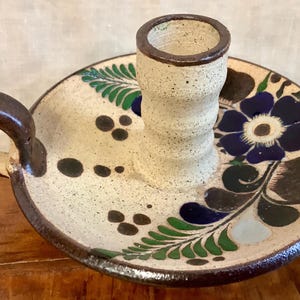 May include: A white ceramic candle holder with a brown handle and a blue and green floral design. The holder has a round base and a tall, cylindrical centre piece.