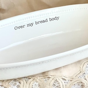 May include: White oval serving dish with handles. The dish has a beaded rim and the words "Over my bread body" printed on the inside. The dish is on a lace tablecloth.