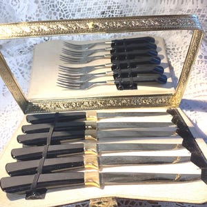 Könnte beinhalten: Ein Set vergoldeter Besteckteile mit schwarzen Griffen in einem vergoldeten Etui. Das Etui ist mit einem cremefarbenen Stoff ausgekleidet. Das Besteck umfasst Gabeln und Messer.