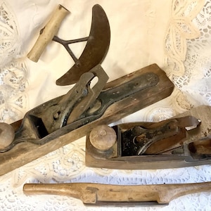 Peut inclure: Ensemble d'outils de menuiserie anciens, comprenant deux rabots en bois, un outil à lame incurvée et une poignée en bois. Les outils sont disposés sur une surface en dentelle blanche, mettant en valeur leur aspect vieilli et leur savoir-faire.