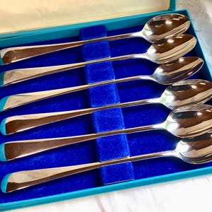 Op de afbeelding: Set van zes zilveren lepels in een blauwe presentatiedoos. De lepels hebben lange, slanke stelen en afgeronde kommen. De doos heeft een blauw fluwelen interieur en een turkooizen rand. De lepels zijn netjes in de doos gerangschikt.