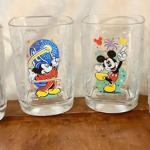 Puede incluir: Un juego de cuatro vasos transparentes con gráficos coloridos de Mickey Mouse en varios atuendos, incluyendo director, hechicero, pose casual y explorador. Cada vaso muestra el logotipo de Disney en la base del diseño impreso.