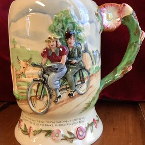 May include: A ceramic mug with a floral design and a handle shaped like a flower. The mug features a scene of a couple riding a tandem bicycle with a cow in the background. The text on the mug reads: "In each trip we take, then if I don't do well,  you to use the brake, my beautiful daisy bell."