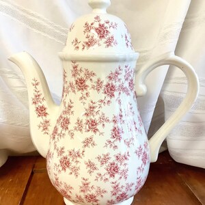 May include: White ceramic coffee pot with a lid and handle. The pot is decorated with a red floral pattern. The spout and handle are curved. The pot is on a wooden surface.
