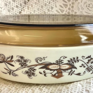 May include: An oval-shaped white casserole dish with a matching translucent amber glass lid. The exterior of the white base features a decorative brown floral pattern with swirling vines and leaves. The dish includes integrated side handles.