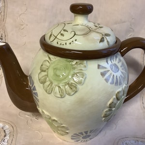 May include: A ceramic teapot with a brown handle and lid. The teapot is decorated with a floral pattern in shades of green, blue, and brown.