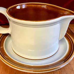 May include: A cream-coloured gravy boat with a brown interior and a matching saucer. The gravy boat has a handle and a spout. The saucer has a brown rim, and the set is made of ceramic.