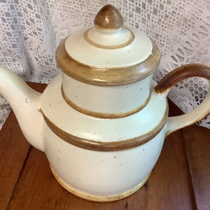 Puede incluir: Una tetera de cerámica blanca con tapa y asa marrones. La tetera tiene un esmalte moteado y bandas marrones alrededor del cuerpo.