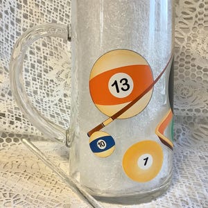 May include: A clear glass beer mug with a handle, featuring a billiard theme. The mug has a white background with a brown pool cue and three billiard balls: a yellow ball with the number 1, a blue and white ball with the number 10, and a white and orange ball with the number 13. A black and white billiard ball with the number 8 is attached to a clear glass stirring rod.