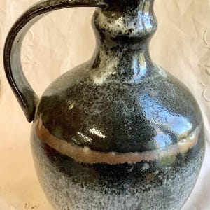 Puede incluir: Jarra de cerámica hecha a mano con cuerpo redondeado y asa curva. La jarra presenta un esmalte oscuro y moteado con una banda marrón y un degradado de gris oscuro a claro en la base. Una pieza única para servir, mide unos 20 cm de altura.