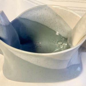 Peut inclure: Un bol en céramique au design unique et fluide. Le bol présente un mélange de teintes bleu clair, blanc et gris. Le bord s'incurve vers le haut, créant une forme distinctive. Le bol est probablement fait à la main.