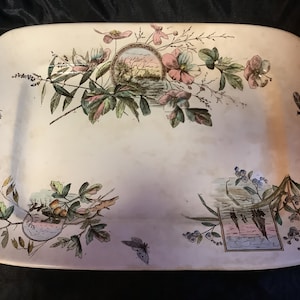 Puede incluir: Un plato rectangular de cerámica blanca con un diseño floral. El plato presenta flores rosas, hojas verdes y mariposas. El centro del plato presenta una escena de paisaje con un río y árboles.