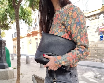 Puede incluir: Un bolso bandolera de cuero negro con una correa larga. El bolso lo lleva una persona que lleva un jersey con estampado floral y unos vaqueros grises.