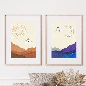 May include: Two framed prints with minimalist illustrations of a sun and moon over a mountain range. The sun print features a gold sun with birds flying above a brown mountain range. The moon print features a silver crescent moon with stars above a blue and purple mountain range.