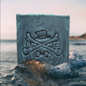 May include: A bar of soap with a green and gray marbled design. The soap features a skull and crossbones with a top hat and bow tie. The text "FB SC" is printed on the soap.