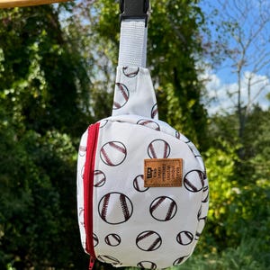 Pode incluir: Bolsa de cintura branca com zíper vermelho e um remendo de couro marrom que diz "I AM KIND SMART IMPORTANT CREATIVE UNIQUE LOVED". A bolsa de cintura é coberta com um padrão de bolas de beisebol vermelhas e brancas.