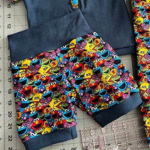 May include: A pair of blue pyjama trousers with a Sesame Street character print. The trousers have a dark blue elastic waistband and cuffs.